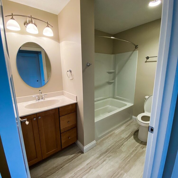 A bathroom featuring a sink with a wooden cabinet, an oval mirror, and wall lights on the left. A bathtub with a curved curtain rod and a toilet are on the right. The floor is tiled.