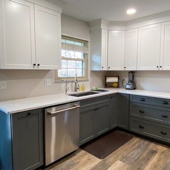 Modern kitchen with white upper cabinets, gray lower cabinets, stainless steel dishwasher, and dark wood flooring. Small window above the sink lets in natural light.