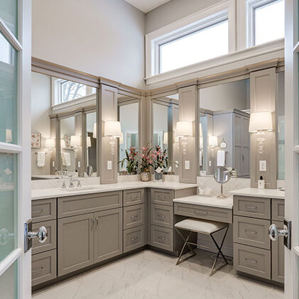 Spacious bathroom with gray cabinets, double sinks, large mirrors, a vanity area with a stool, white countertops, and tall windows providing natural light—see our pricing guide to help plan your renovation.